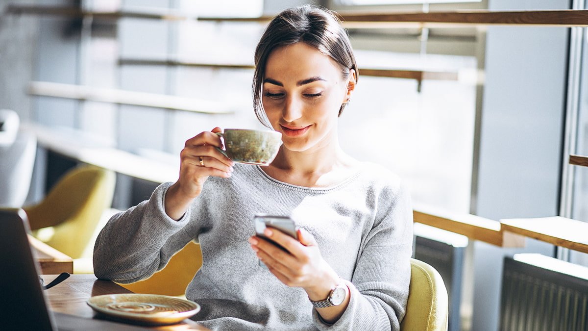 marketing de defensores - cliente sorrindo segurando uma xícara de café sentada em uma poltrona e utilizando o celular
